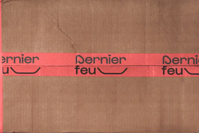 Un carton brun fermé par une bande adhésive rouge sur laquelle le logotype Dernier feu est imprimé en noir, répété sur toute la longueur.